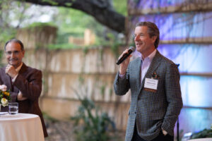 Man in checked jacket holding microphone speaks at event in a botanical garden setting