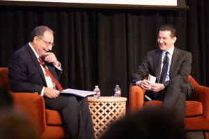Two men wearing dark suits laugh together as they sit in red chairs on a stage