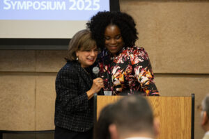 Two women embrace at podium in front of large screen while one holds microphone and speaks to audience