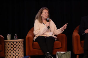 Woman with long brown hair wearing pale pink top holds microphone to address audience
