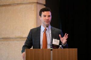 Man in dark jacket and red tie speaks into microphone from podium