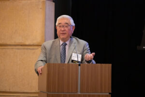 Man white hair and glasses wearing pale gray jacket and blue tie speaks from a podium