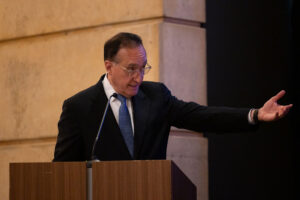 Man wearing dark suit and blue tie stands at podium and gestures at audience