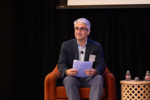 Man with white hair and glasses wearing a dark jacket holds papers as he sits in red chair
