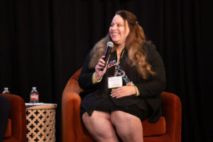 Woman with long brown hair wearing black suit sits in red chair holding microphone and smiling