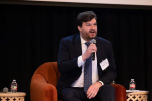 Man with dark brown hair and beard speaks into microphone