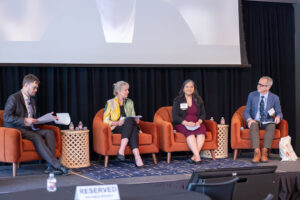 Two men and two women sit in red chairs on stage in front of a large screen