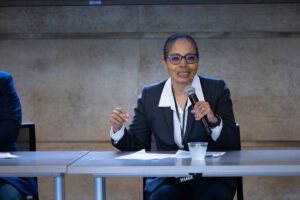 Woman wearing white shirt, black blazer and glasses holds microphone to address audience