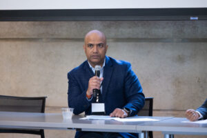 Man wearing blue blazer sits at table holding microphone to address audience