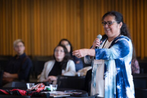Woman wearing glasses and a blue and white top speaks to audience using microphone.