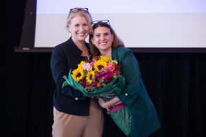 Description: Two women embrace while holding a bouquet of bright flowers. Context: Sara Wise and Donna Musselman at GHS 2025