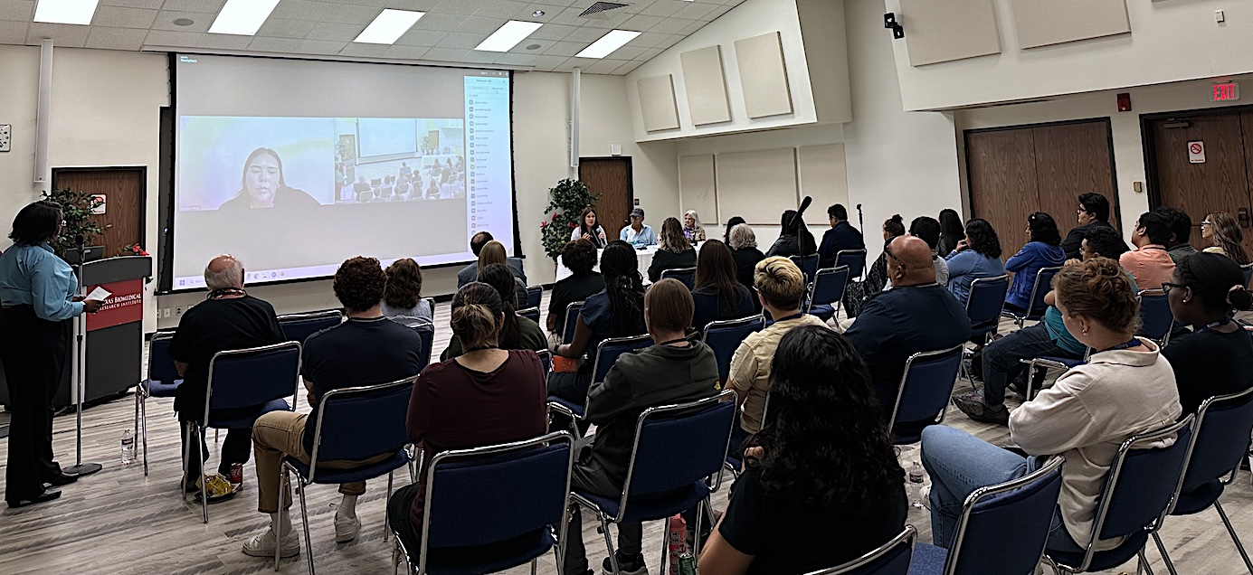 Conference room with people sitting in chairs looking at large screen and listening to speaker at the front of the room.