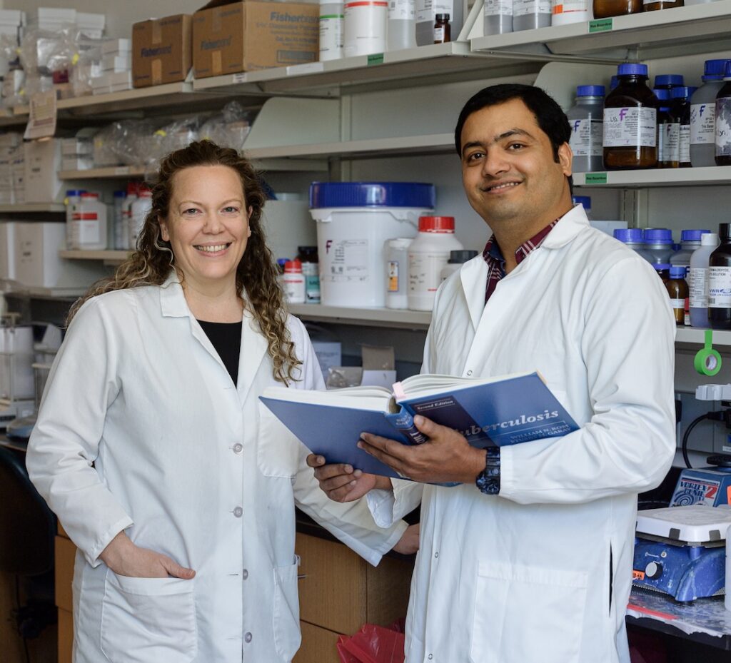 A female and male scientist stand in a research lab, smiling at the camera.