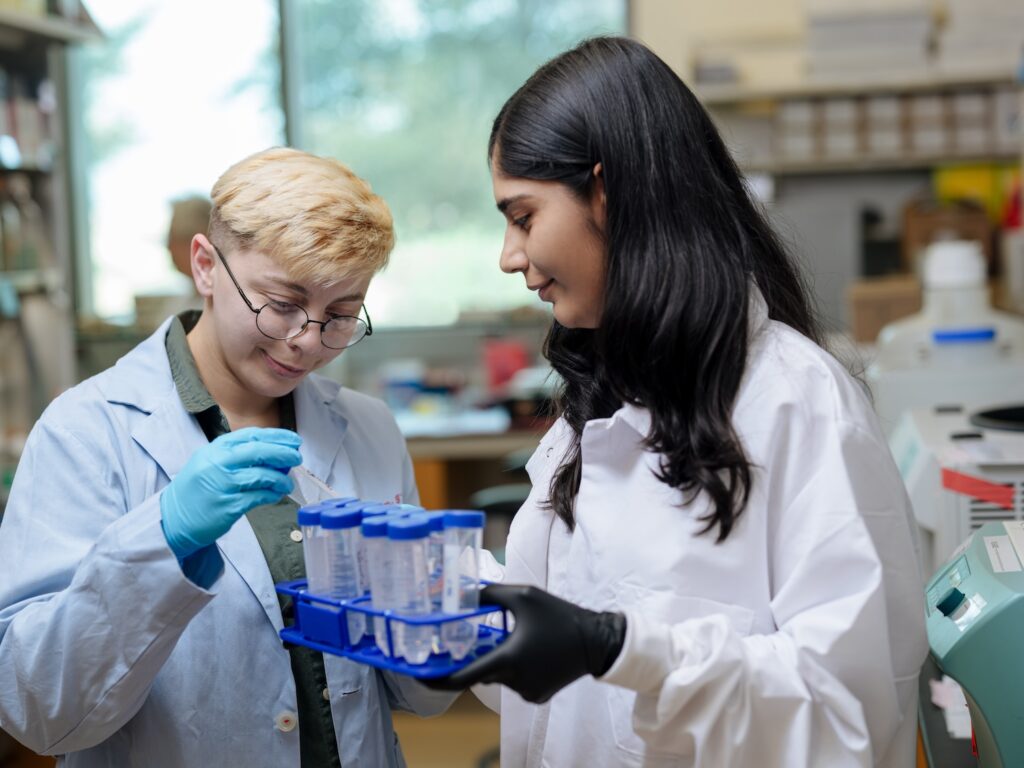 Two young women in white lab coats in a lab and wearing latex gloves hold a tray of plastic tubes.