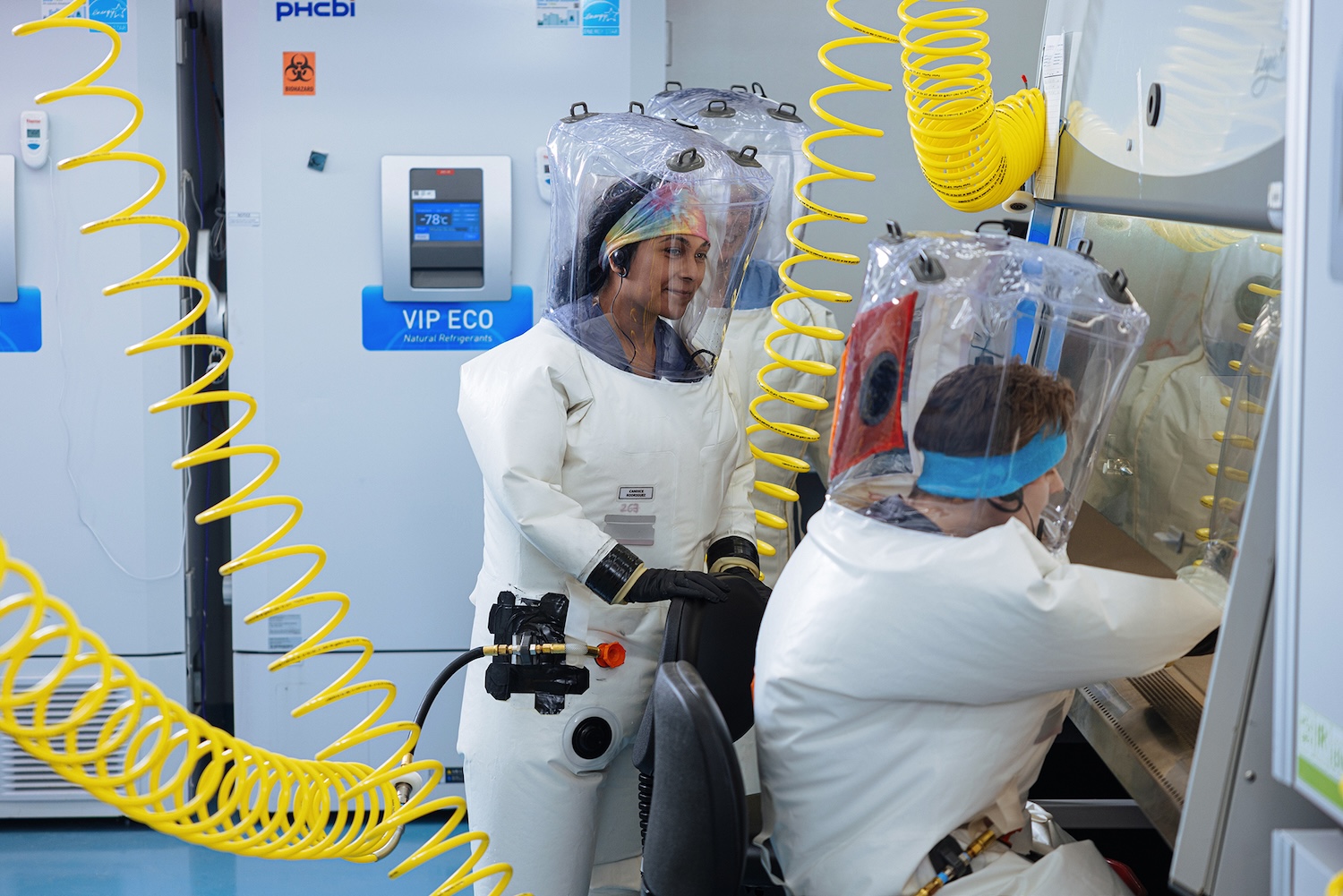 Researchers in full body suits working inside a BSL-4 lab. Yellow coiled air hoses stretch from the ceiling to the inflated suits.