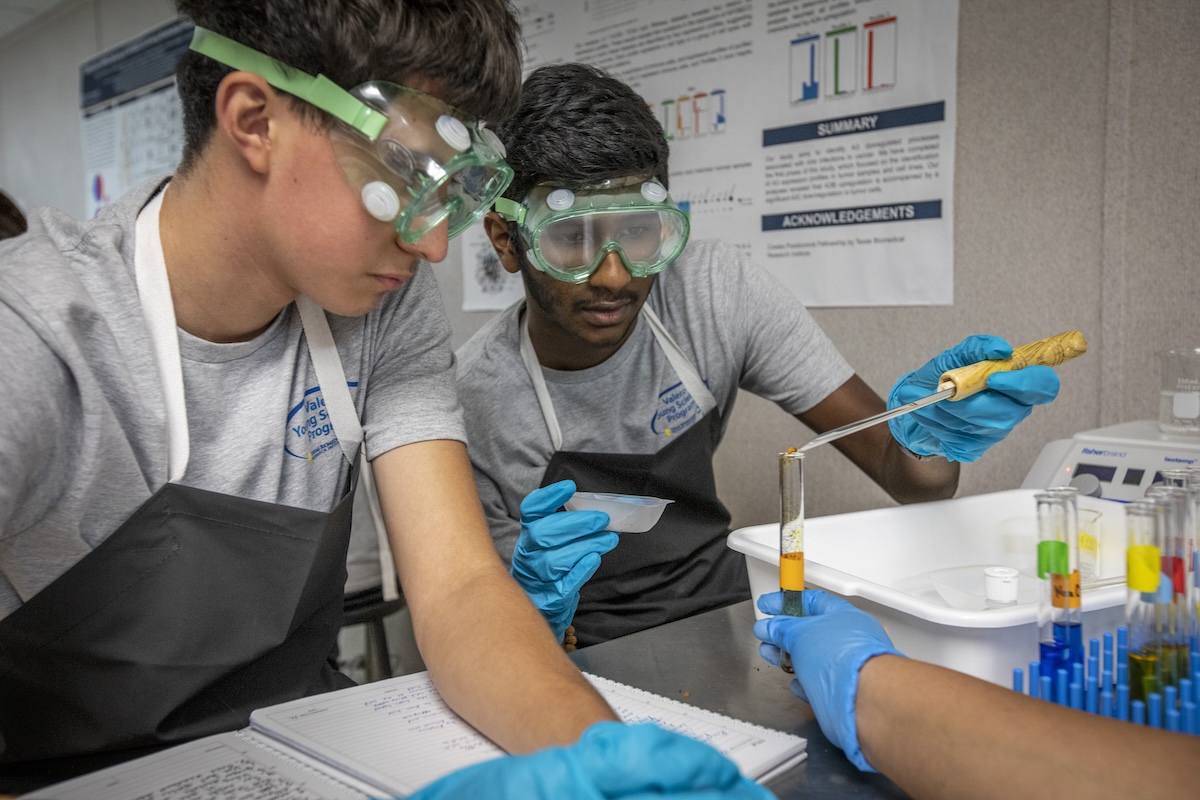 One student adds to a test tube being held by another as a third student looks on One student adds to a test tube being held by another as a third student looks on